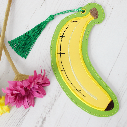 Banana-shaped bookmark with a green border on a light wooden surface with a pink flower.