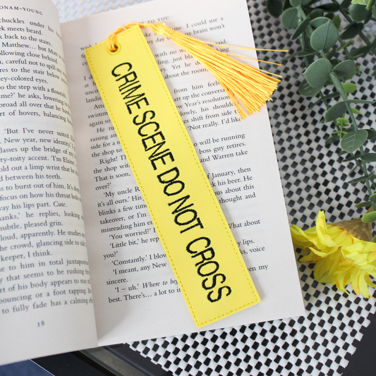 Yellow 'CRIME SCENE DO NOT CROSS' bookmark on an open book with a tassel.