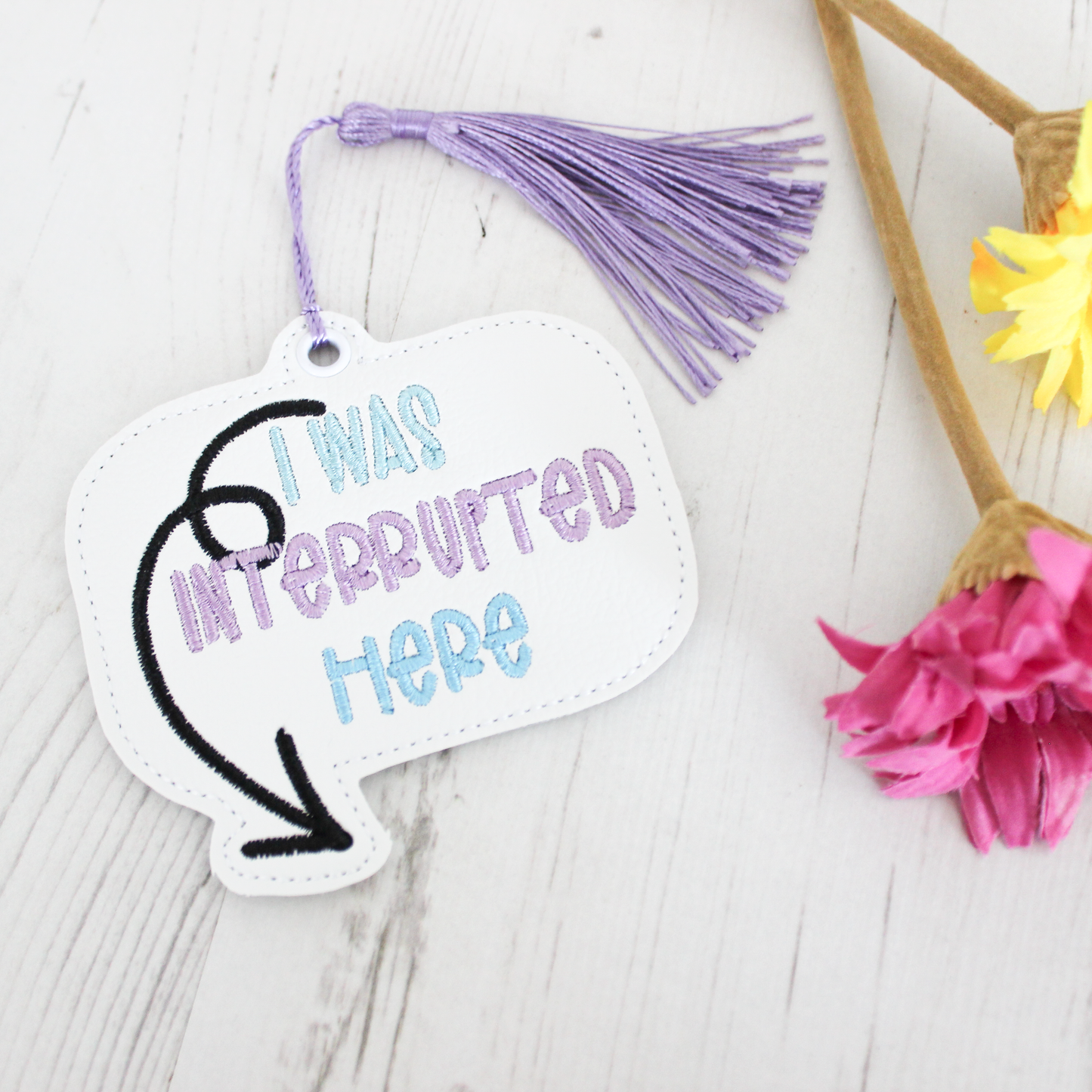 Bookmark with 'I was interrupted here' text on a white surface with flowers.