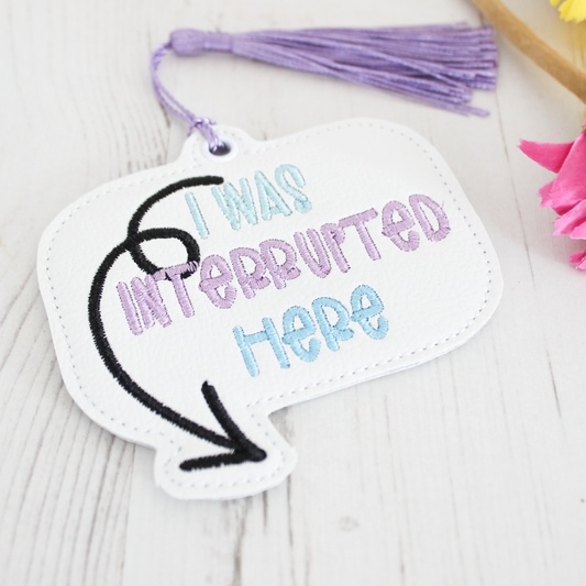 White sign with 'I was interrupted here' text and purple tassel on a light wooden surface.
