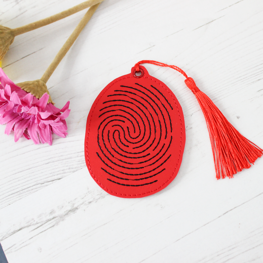 Red finger print suspect novelty bookmark with a tassel on a white surface with pink flowers and dried plants.
