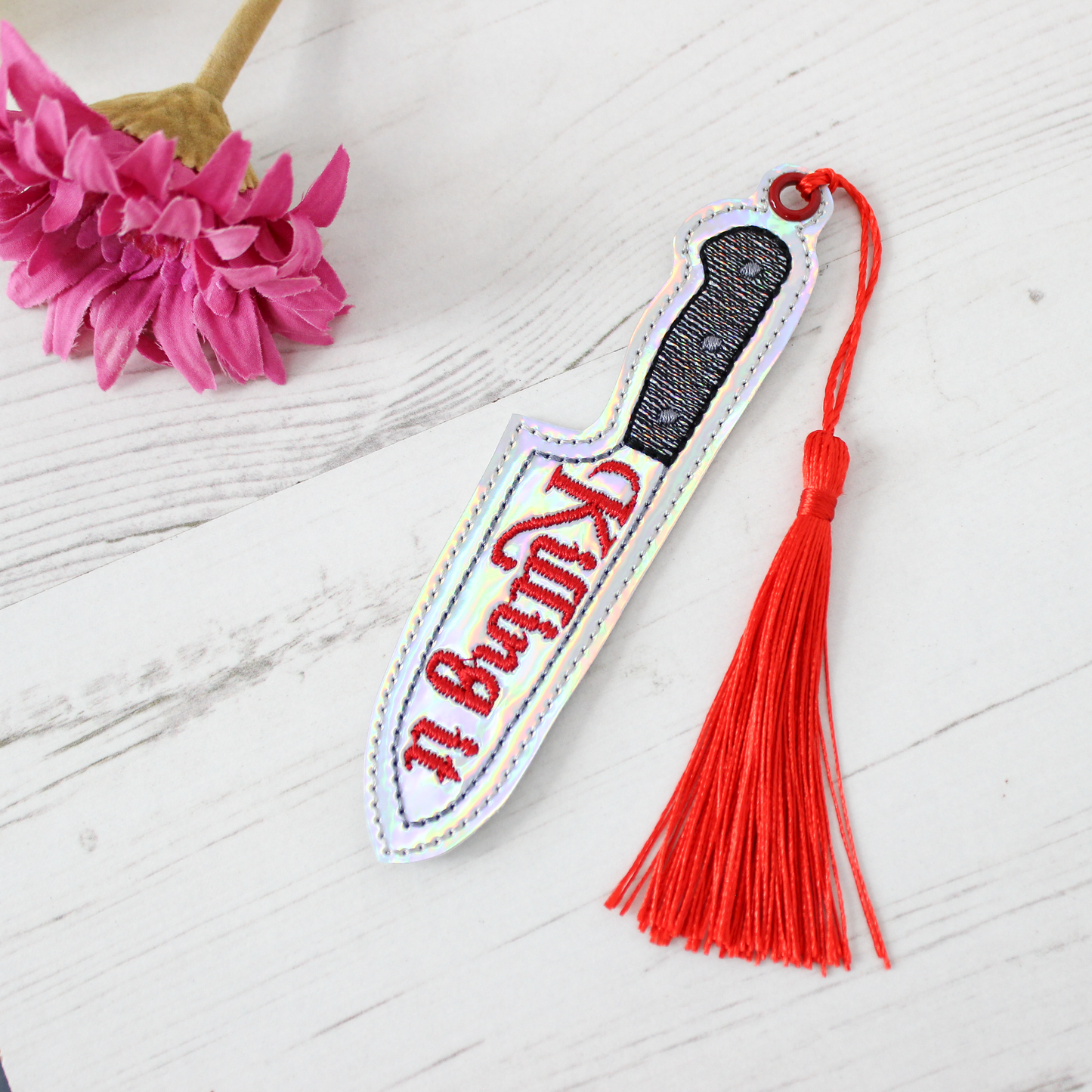 Decorative knife-shaped keychain with red tassel on a white surface with pink flowers.