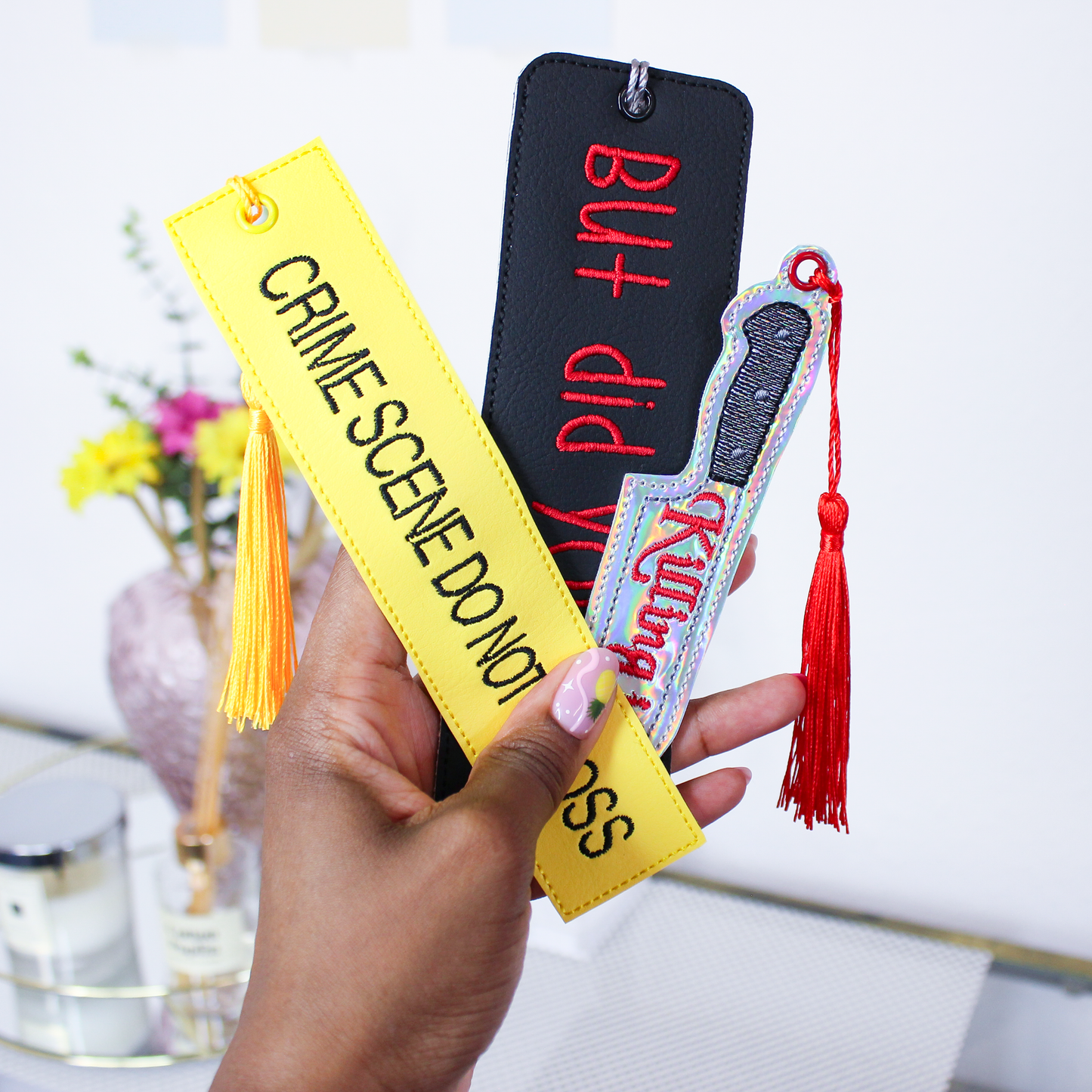 Hand holding three unique true crime themed bookmarks with tassels against a blurred indoor background