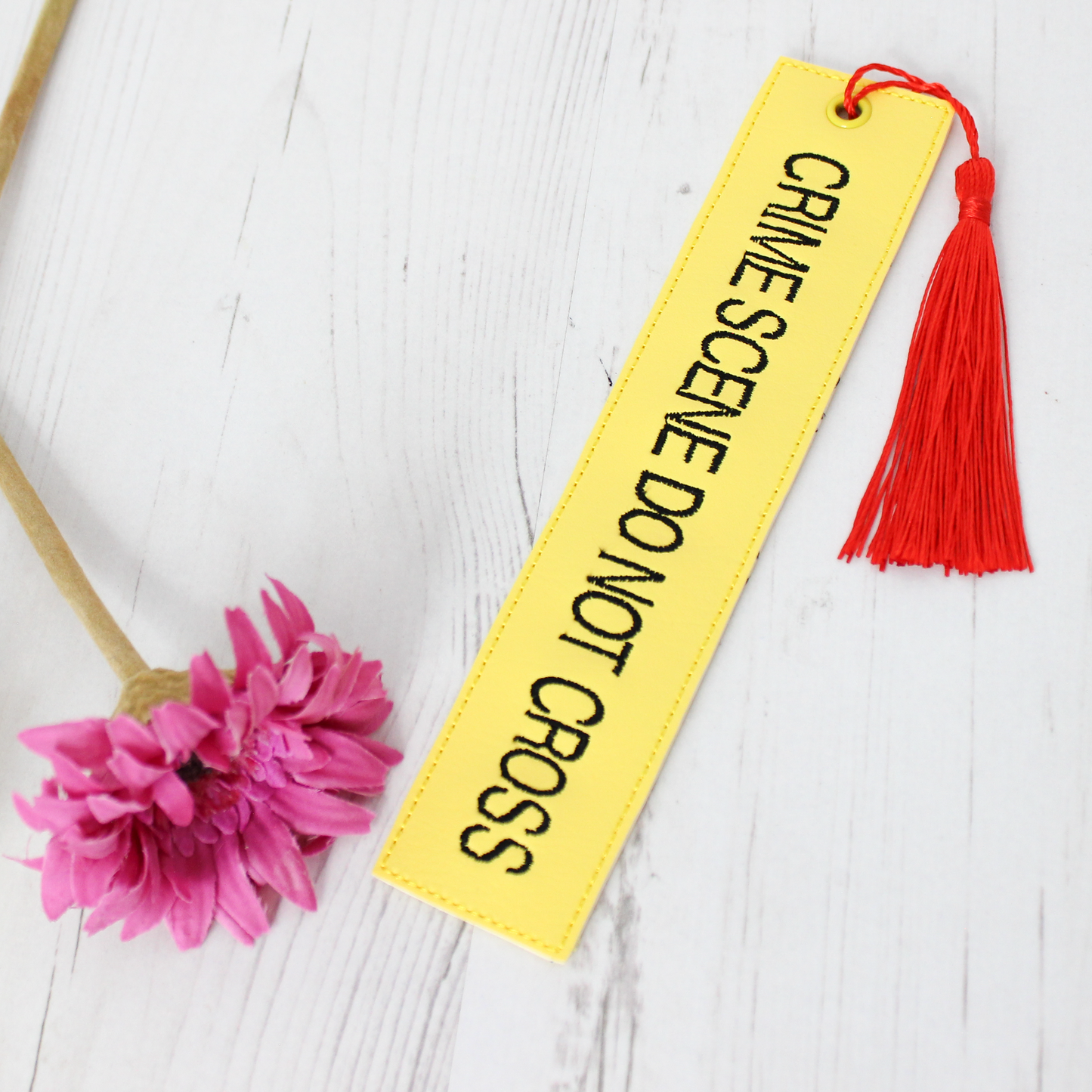 Yellow bookmark with 'CRIME SCENE DO NOT CROSS' text and red tassel on a white wooden surface with a pink flower.