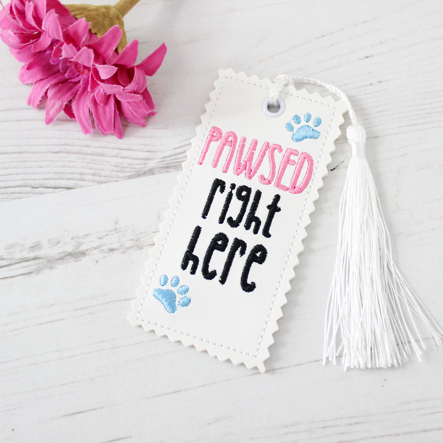 Bookmark with 'PAWSED right HERE' text and tassel on a light wooden surface with pink flowers.