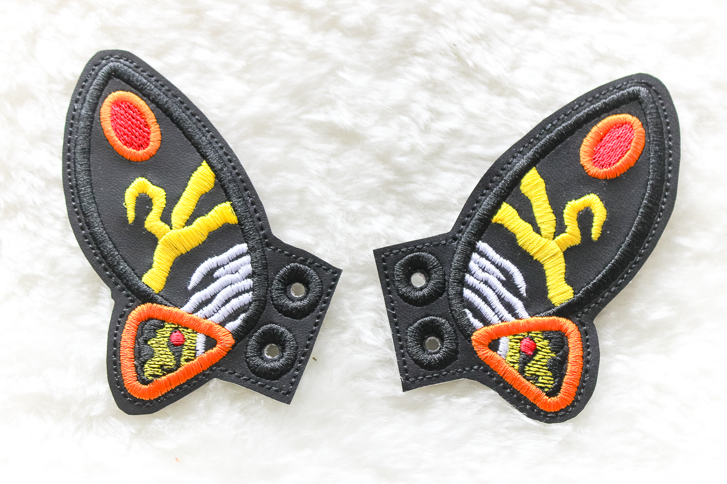 A pair of MOTHRA cosplay shoe wings with a central emblem on a white textured background
