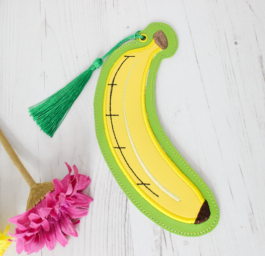 Banana-shaped bookmark with green tassel on a light wooden surface with pink flower