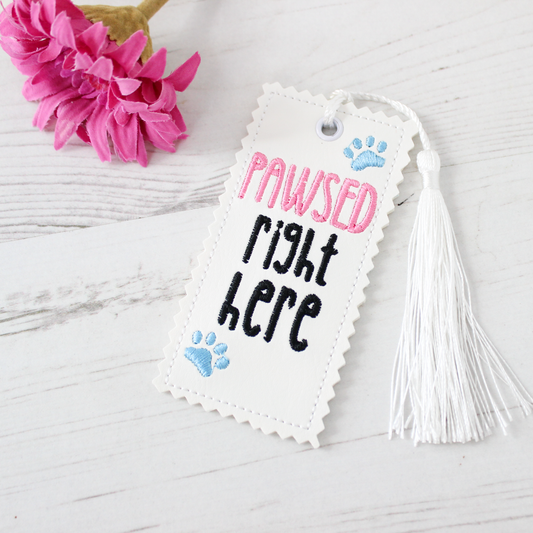 Bookmark with 'PAWSED right HERE' text and tassel on a light wooden surface with pink flowers.